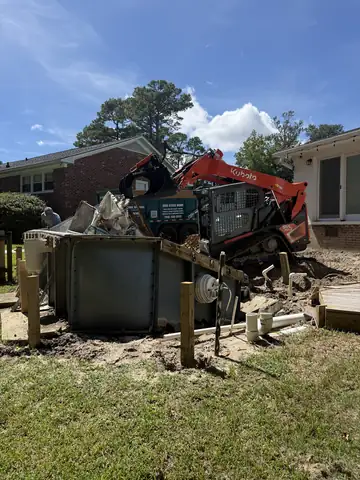 Kubota track loader breaking down above-ground pool walls beside a brick house