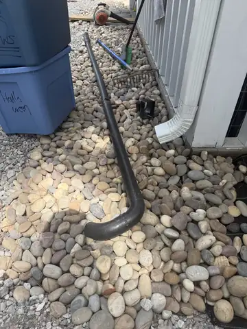 Downspout drain pipe running through a river rock bed beside a house