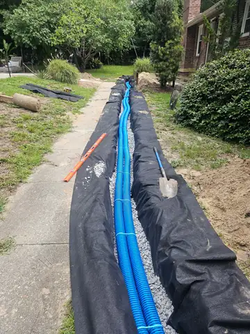 French drain trench lined with fabric and blue perforated pipe beside a walkway